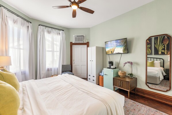 A bedroom with a bed, a ceiling fan, a TV, a wardrobe, a small cabinet, and a mirror.