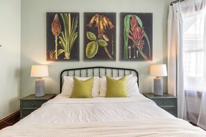 A neatly made bed with white bedding, two green pillows, and a black headboard. There are two bedside tables with lamps and a wall-mounted painting of plants.