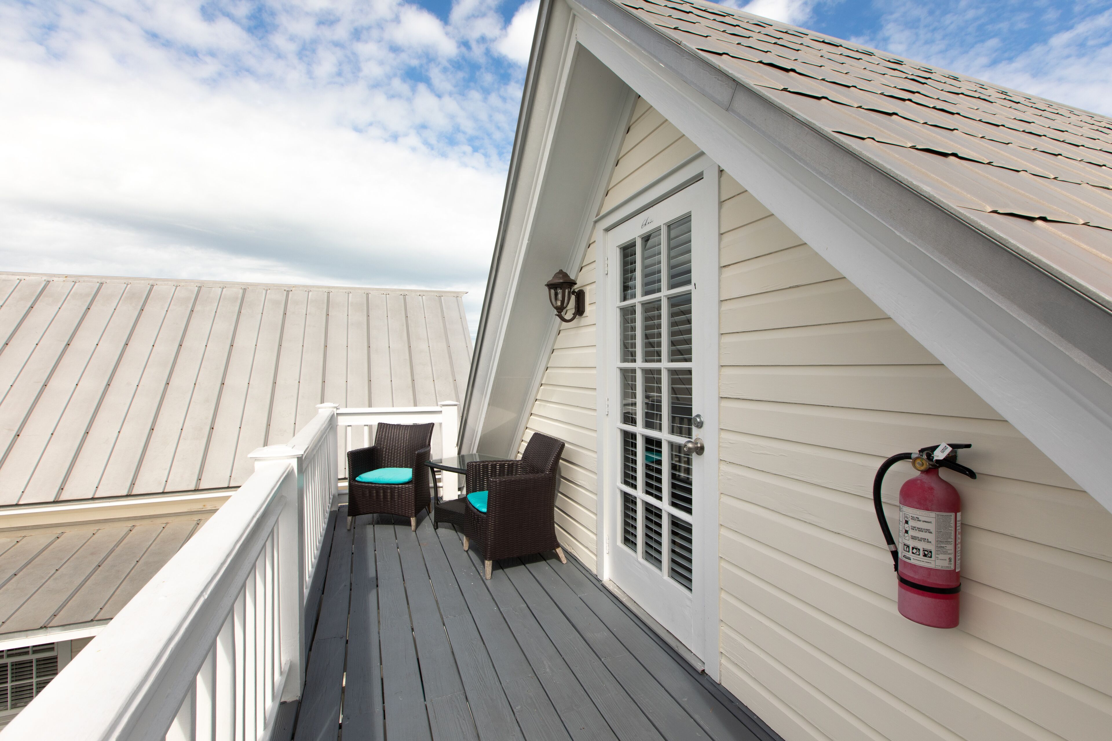 A deck with two wicker chairs and a table, a fire extinguisher mounted on the wall, and a white door with a window.
