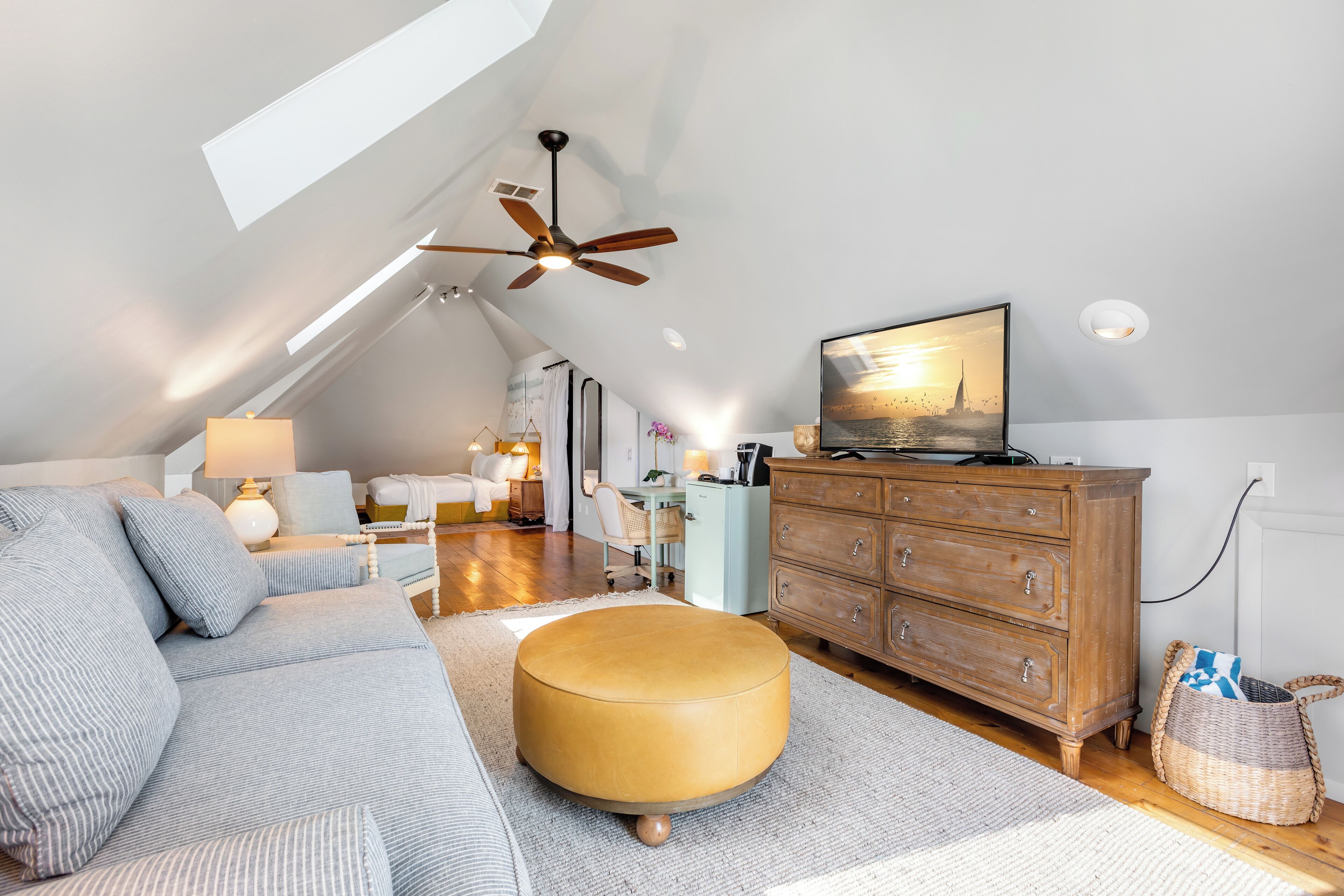 Une chambre de style loft moderne avec un plafond en pente, un ventilateur de plafond, une grande fenêtre, un téléviseur, une commode en bois, un canapé et un pouf rond.