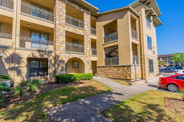 A multi-story building with balconies, stone accents, and a parking area with cars.