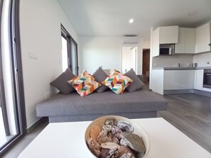 A modern living room with a grey sofa, colorful geometric cushions, a wooden side table with seashells, and a kitchen in the background.