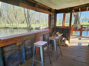 A wooden cabin with a deck overlooking a pond, two metal stools, and a stack of firewood. - Creek Side Cottage with Lake Views, Lake Access, Dock and Secluded Yard (Jamestown)