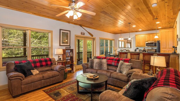 A spacious living room with wooden ceilings, a ceiling fan, and a large window overlooking greenery. - Rivers Rest – Beautiful Riverfront Townhome in Downtown Helen (Helen)