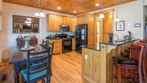 A spacious kitchen with wooden cabinets, a central island, and a dining area with chairs. - Rivers Rest – Beautiful Riverfront Townhome in Downtown Helen (Helen)