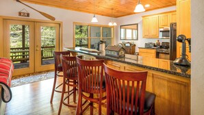 A kitchen with wooden cabinets, a granite countertop, and a dining area with wooden chairs. - Rivers Rest – Beautiful Riverfront Townhome in Downtown Helen (Helen)