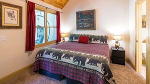 A bedroom with a wooden ceiling, a large window with red curtains, a bed with a patterned bedspread, two bedside lamps, and a painting on the wall. - Rivers Rest – Beautiful Riverfront Townhome in Downtown Helen (Helen)