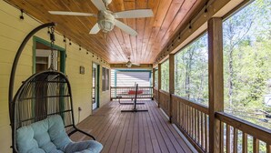 A wooden deck with a ceiling fan, a wicker chair, and a view of the forest. - Rivers Rest – Beautiful Riverfront Townhome in Downtown Helen (Helen)
