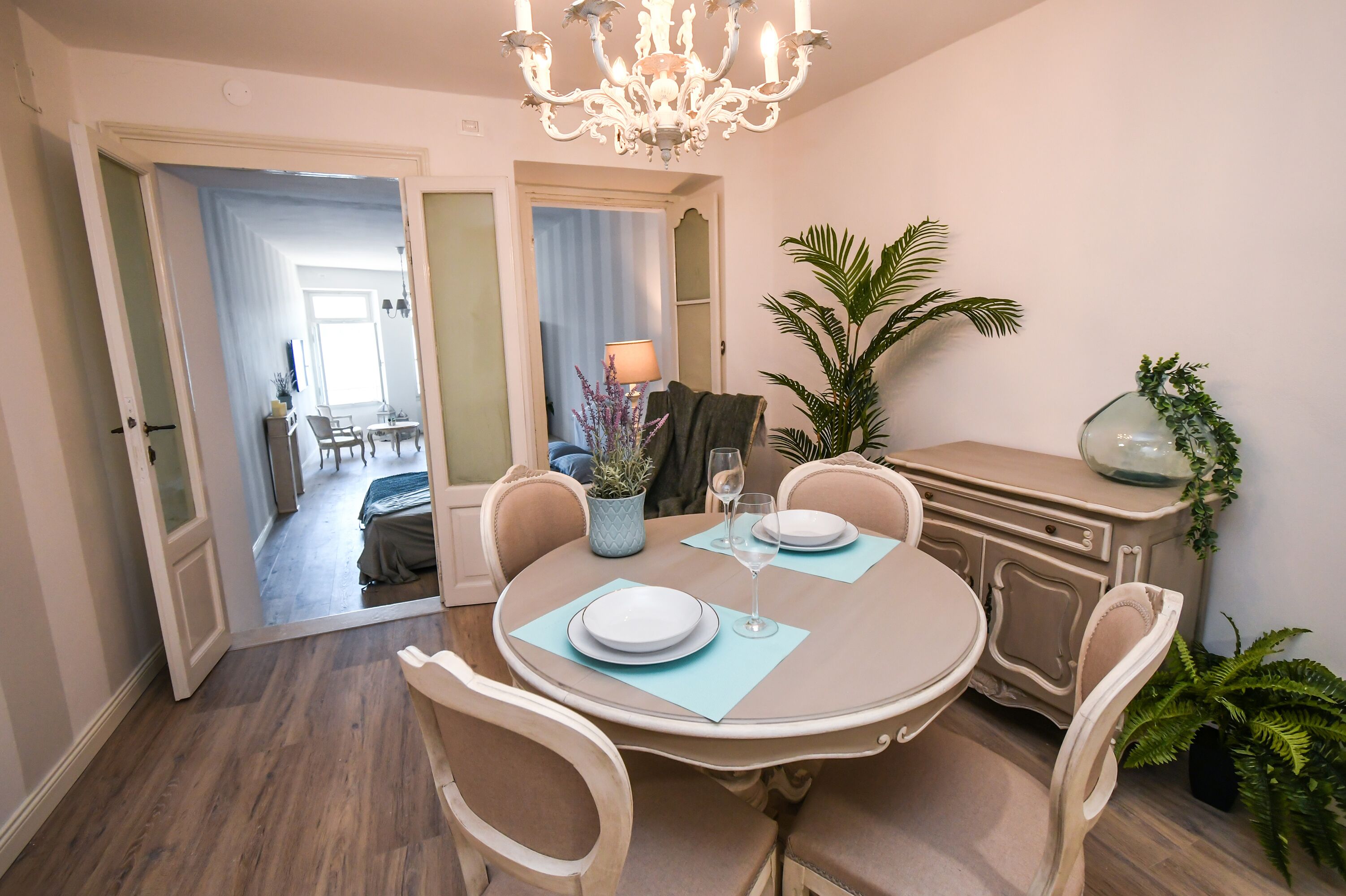 A dining area with a round table set for two, chairs, a chandelier, and a potted plant.
