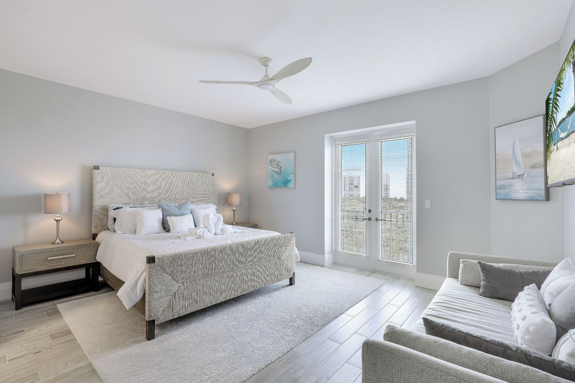 A bedroom with a bed, bedside tables, a ceiling fan, a door with blinds, and a painting on the wall.