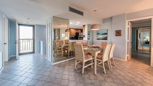 A modern interior with a dining area featuring a glass table and wicker chairs, a kitchen with dark cabinets, and a balcony with a view. - Surf Colony - Vanderbilt Beach - Bayside (Naples)
