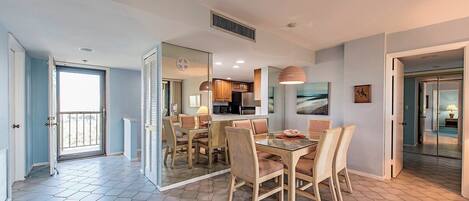 A modern interior with a dining area featuring a glass table and wicker chairs, a kitchen with dark cabinets, and a balcony with a view.