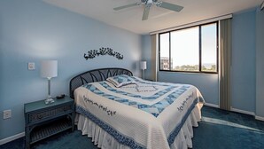 A bedroom with a bed, a nightstand, a lamp, a ceiling fan, and a window with curtains. - Surf Colony - Vanderbilt Beach - Bayside (Naples)