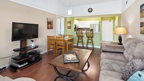 A living room with a flat-screen TV, wooden dining table, and chairs, a sofa, and a kitchen in the background. - Key Royal - William Raveis Rentals (Naples)
