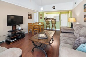 A living room with a flat-screen TV, wooden dining table, and chairs, a sofa, and a kitchen in the background. - Key Royal - William Raveis Rentals (Naples)