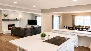 A modern kitchen with a central island, a dining area with a table and chairs, a living room with a sofa and fireplace, and a TV mounted on the wall.