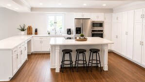 A modern kitchen with white cabinetry, a central island with stools, stainless steel appliances, and a window providing natural light.