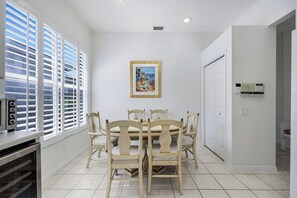 A dining area with a round table and chairs, a painting on the wall, and a window with blinds. - Stonebridge Country Club (Naples)