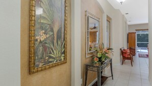 A hallway with a large tropical-themed painting, a glass console table with a floral arrangement, and a wooden chair. - Stonebridge Country Club (Naples)
