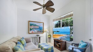 A living room with a sofa, ottoman, and chair, a television, a ceiling fan, a painting, and a window with a beach view. - Stonebridge Country Club (Naples)