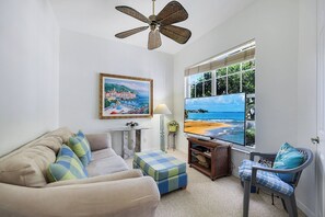 A living room with a sofa, ottoman, and chair, a television, a ceiling fan, a painting, and a window with a beach view.