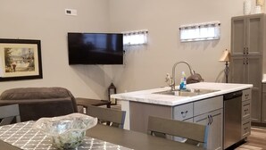 Una cocina compacta con lavavajillas integrado, un pequeño comedor con mesa y sillas, un televisor montado en la pared y un cuadro enmarcado.
