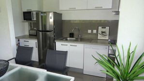 A compact kitchen with a microwave, refrigerator, sink, and dishwasher. There are two chairs and a table with a glass top. A potted plant is visible in the corner. - LE FLAMBOYANT, triplex sea view, 3 bedrooms, residence with swimming pool (le Gosier)
