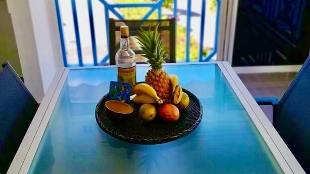 A table with a tropical fruit platter, a bottle, and a card on a glass table with a view of the ocean and a blue railing. - LE FLAMBOYANT, triplex sea view, 3 bedrooms, residence with swimming pool (le Gosier)