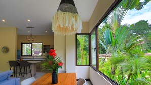 A modern dining area with a wooden table, chairs, and a hanging light fixture. A large window offers a view of lush greenery outside. - Macaws and Sloths and Monkeys, Oh My! Villa Capuchin in Manuel Antonio (Manuel Antonio)