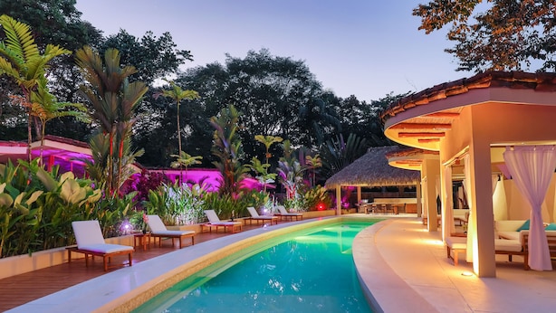 A swimming pool with lounge chairs, surrounded by tropical plants and a thatched-roof structure. - Macaws and Sloths and Monkeys, Oh My! Villa Capuchin in Manuel Antonio (Manuel Antonio)