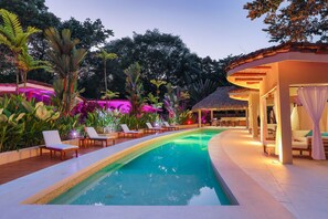 Ein Swimmingpool mit Liegestühlen, umgeben von tropischer Vegetation und einer Struktur mit Reetdach.