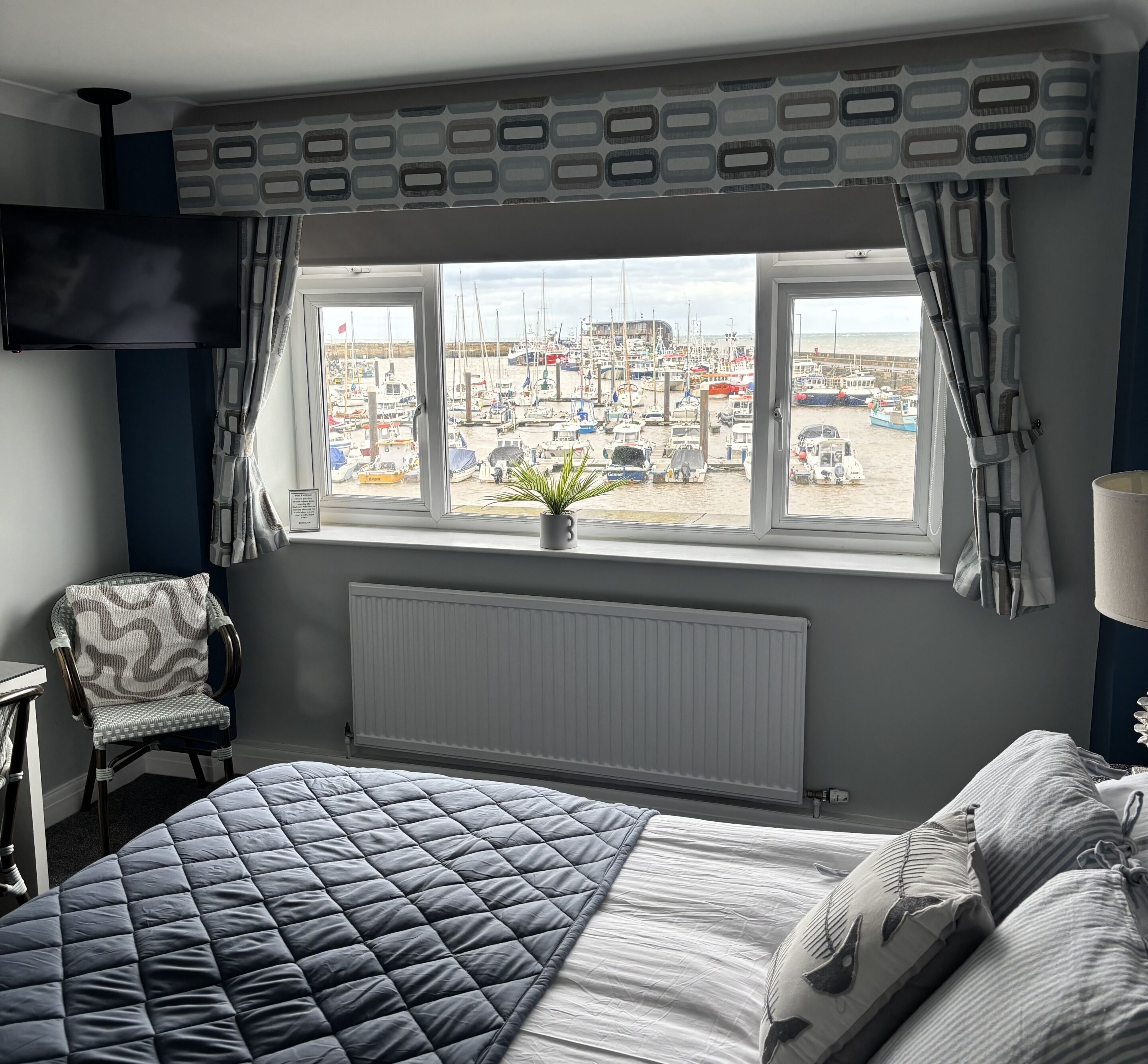 A bedroom with a large bed, a chair, a window with a view of boats and a pier, and a radiator.