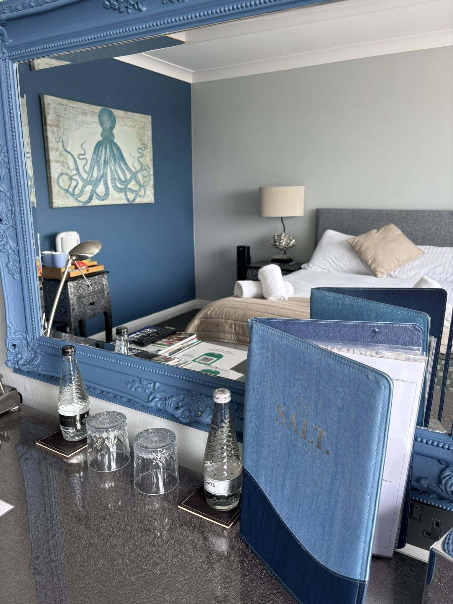 A hotel room with a bed, a bedside table with a lamp, a mirror, and a painting on the wall.