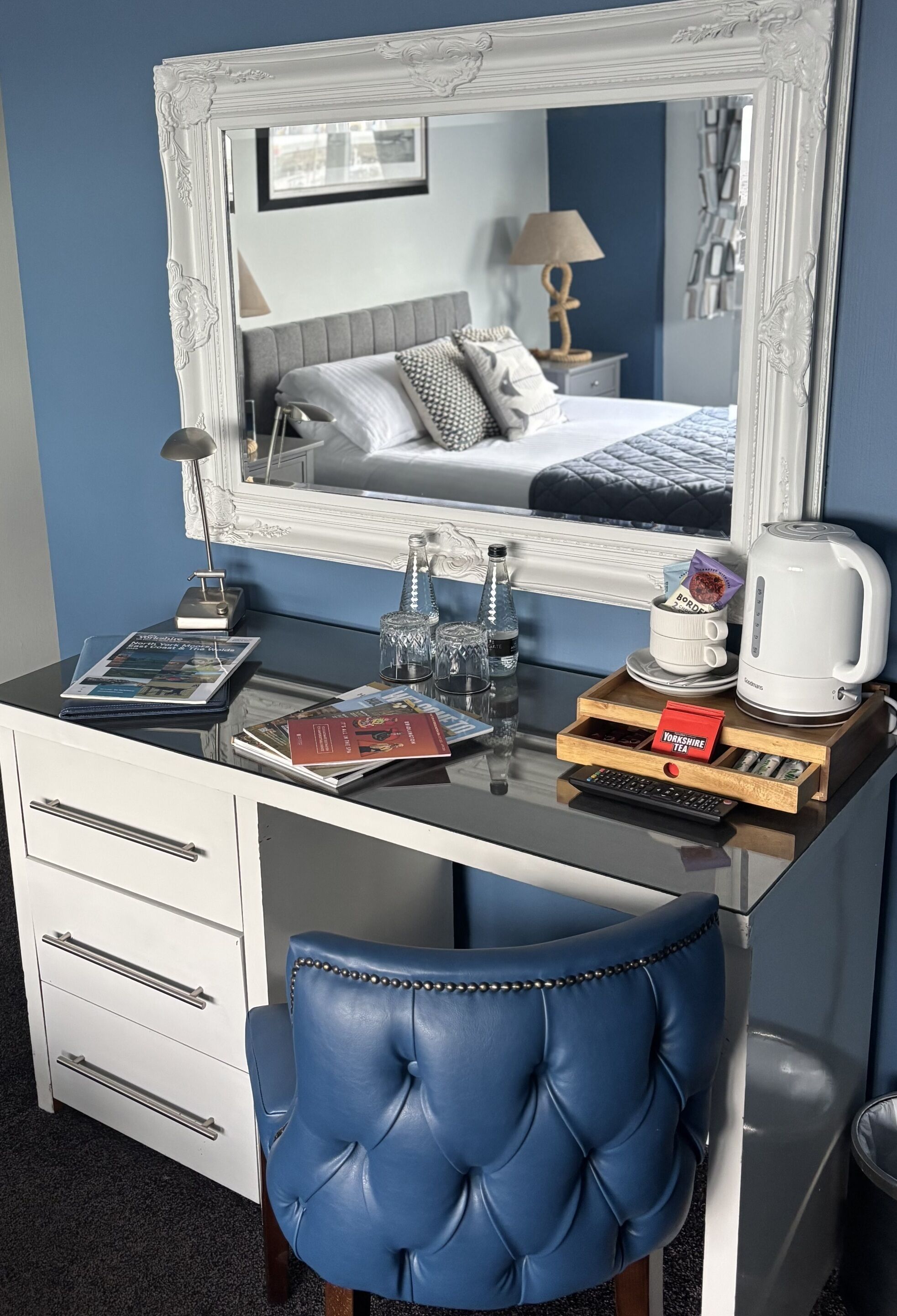 Una habitación de hotel con un espejo grande, una cama con almohadas, una mesita de noche con lámpara, una cafetera y una silla azul.