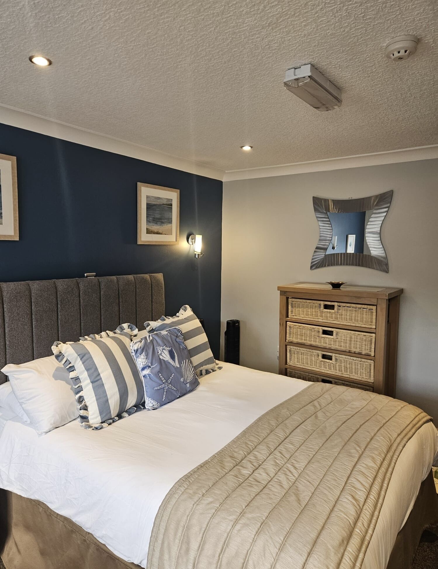 A bedroom with a bed, a headboard, a bedside table with a lamp, a mirror, and a wall-mounted light fixture.
