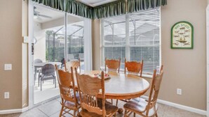 Un coin repas avec une table ronde et six chaises en bois, une porte vitrée donnant sur l’extérieur et une horloge fixée au mur.