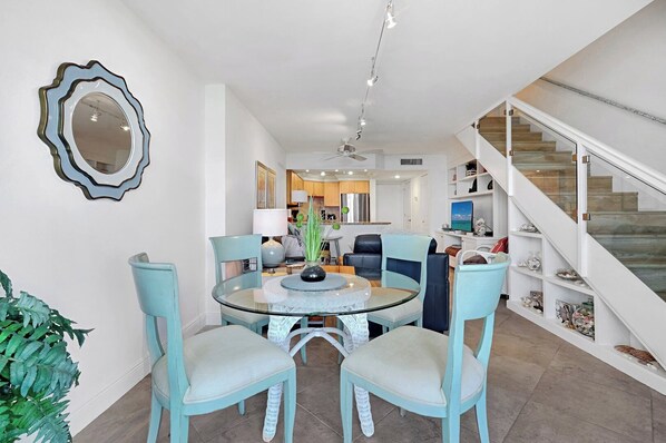 Un interior moderno con un comedor que incluye una mesa y sillas de vidrio, una cocina al fondo y una escalera que sube a otro nivel.