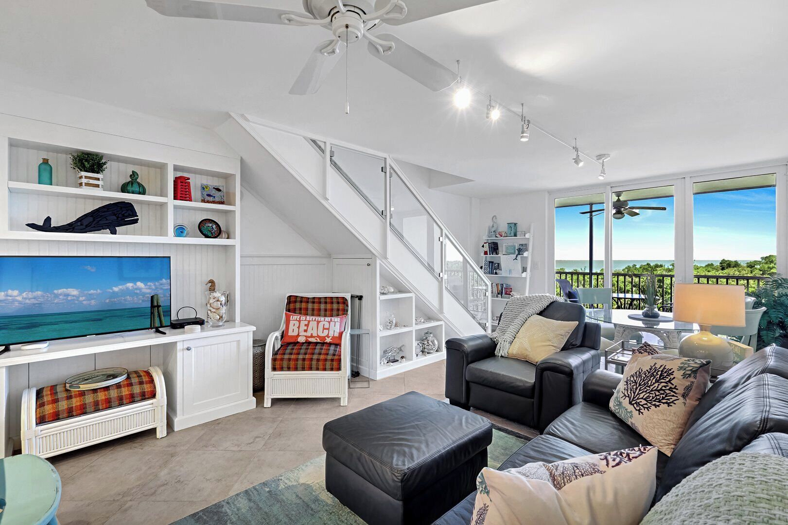 A spacious living room with a large flat-screen TV, a comfortable seating area with a sofa and chairs, a staircase leading to an upper level, and a balcony with a view.