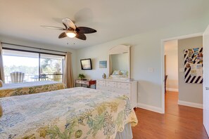 Quarto com cama grande, cômoda, ventilador de teto e janela com vista para árvores.