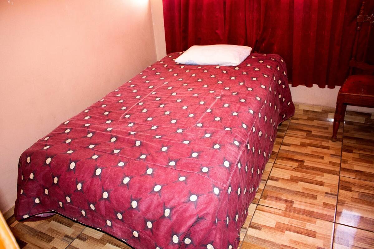 A single bed with a red polka dot bedspread and a white pillow, wooden flooring, and a red curtain.