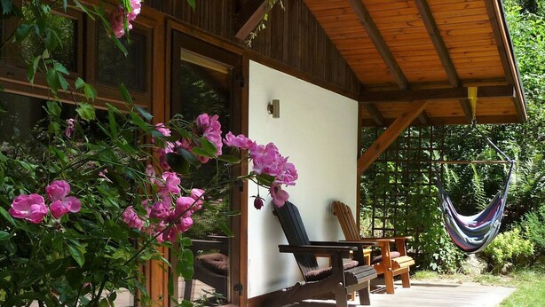 Une terrasse en bois avec deux chaises longues et un hamac, entourée d’une végétation luxuriante et de plantes en fleurs.