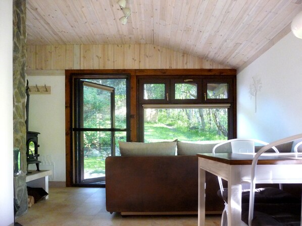 A cozy room with a wooden ceiling, a large window with a view of greenery, a brown sofa, and a white table.