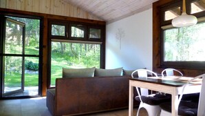 Une pièce chaleureuse avec un plafond en bois, un canapé agrémenté de coussins, une table à manger avec des chaises, et de grandes fenêtres donnant sur une zone boisée.