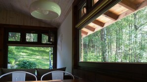Un coin repas avec une table en bois et des chaises blanches, de grandes fenêtres donnant sur la forêt et un ventilateur de plafond.