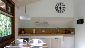 Une cuisine avec une table en bois, des chaises blanches, une lampe suspendue, une horloge et une fenêtre donnant sur des arbres.