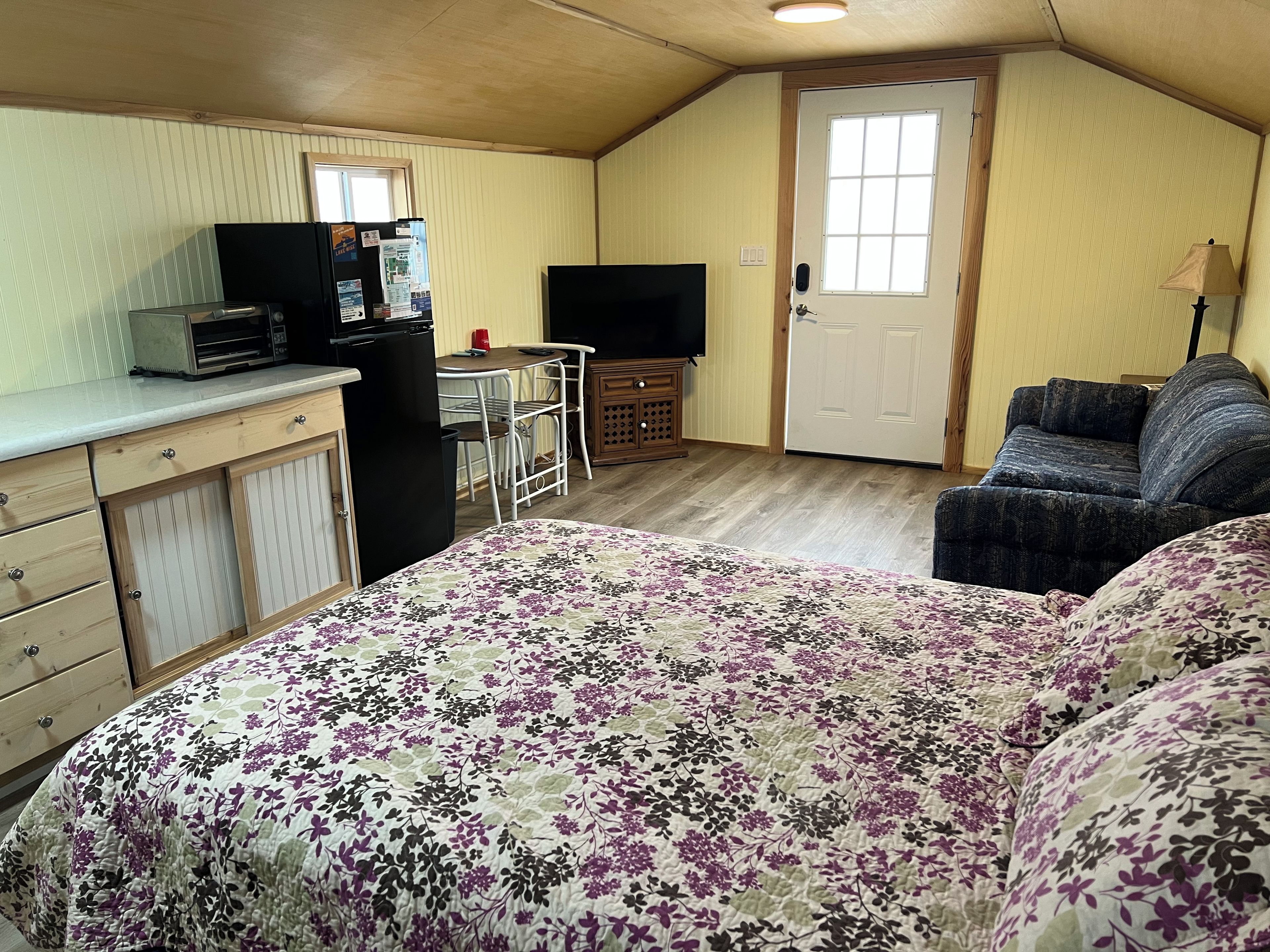 A compact room with a bed, a kitchenette, a television, and a door.