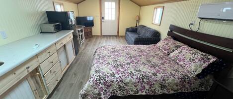 A compact room with a bed, a kitchenette, a television, and a window.