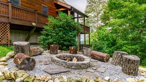 Un foyer en pierre, entouré de rochers et de bûches, avec une terrasse en bois et une maison en arrière-plan.