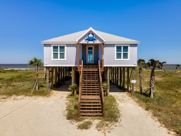 A house on stilts with a front porch and a staircase leading to the entrance. - Bayfront with Private Pool! Outdoor games, modern style! - The Blue Crab (Dauphin Island)