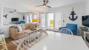 A modern living room with a white sofa, a dining table, and a ceiling fan. - Enjoy Sunset Views at Cozy Oceanfront Condo + Pool on Ocean Isle Beach, NC (Ocean Isle Beach)